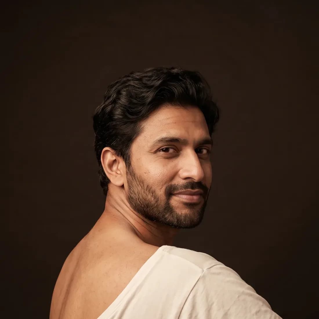 A man seen from behind looking over his bare shoulder, on a deep brown background