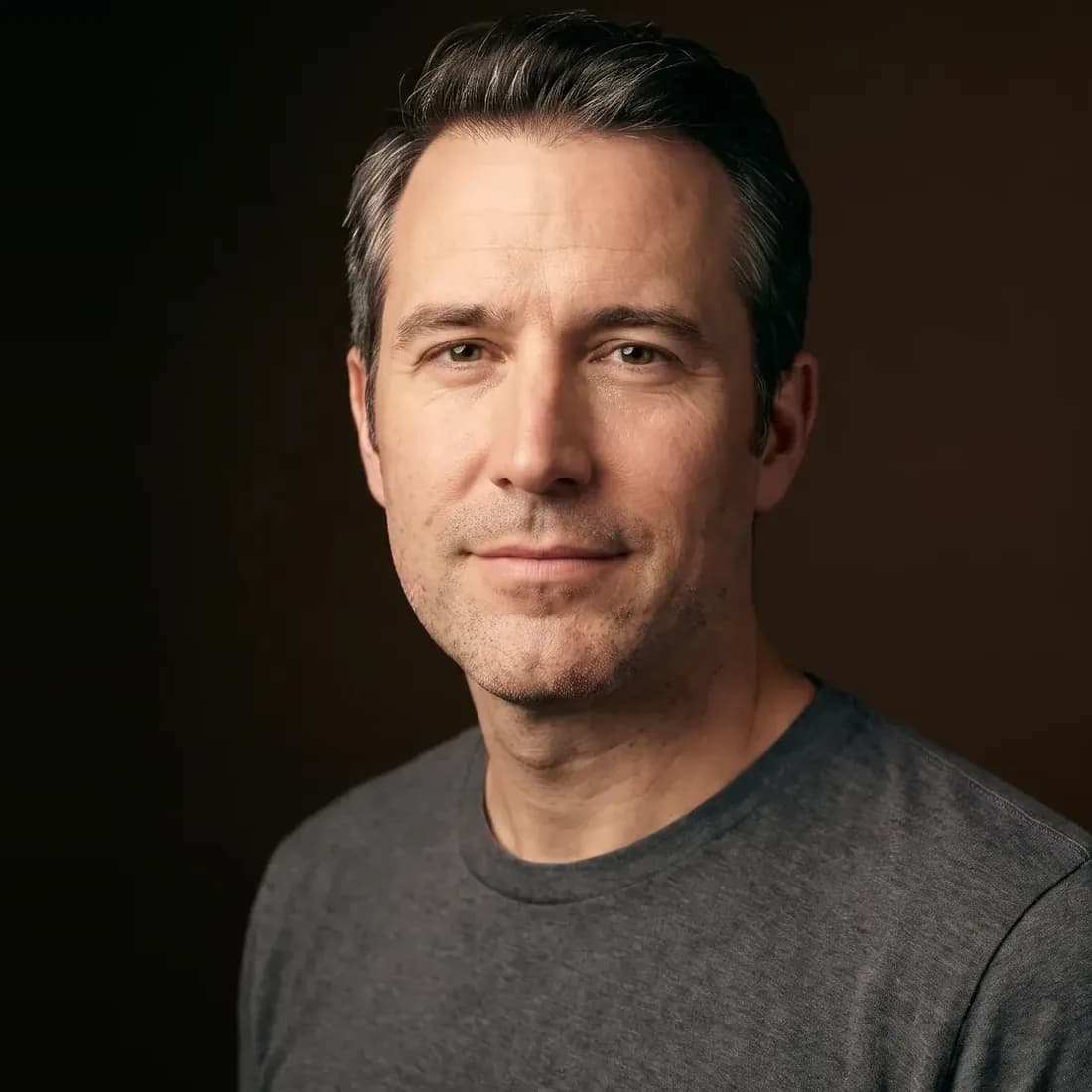 A man with greying temples, closely shaved, holding back a smile, on a deep brown background