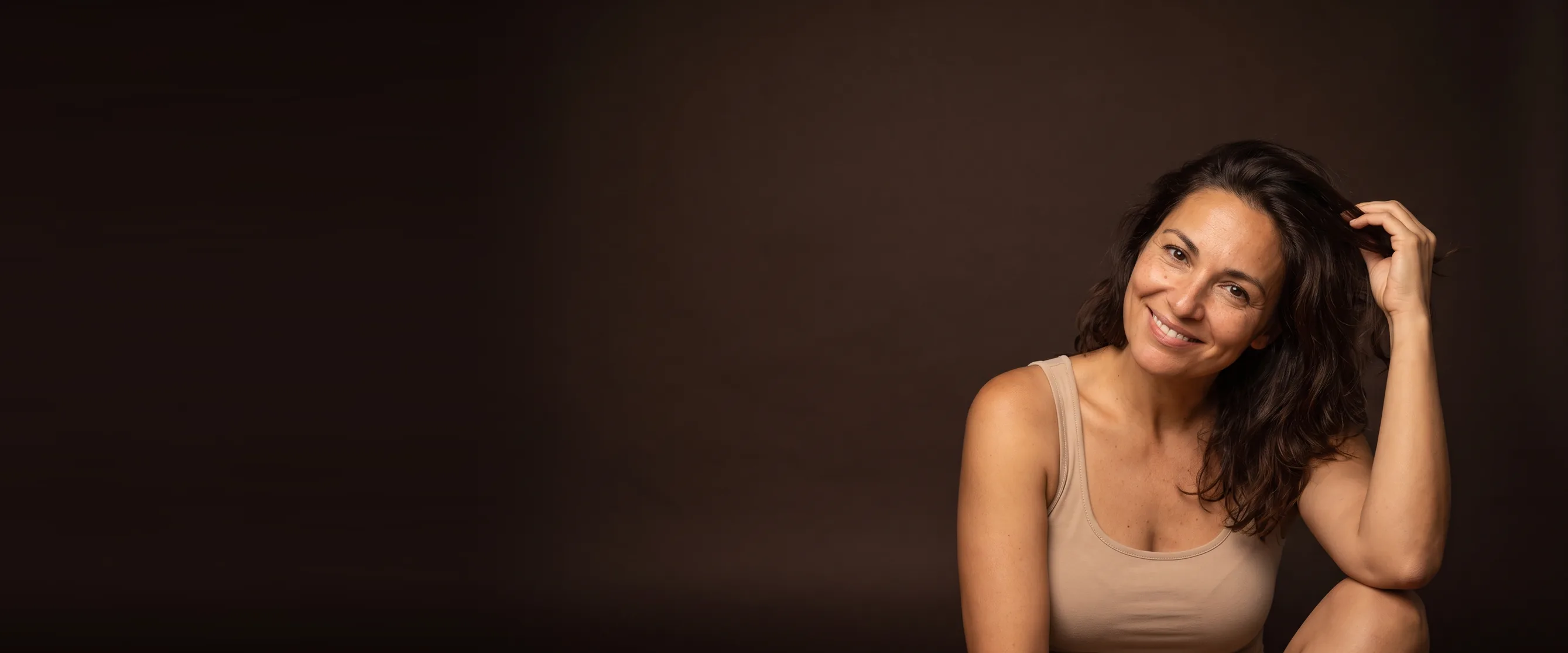 Femme aux cheveux bruns fins, une main soulevant ses cheveux depuis la tempe, sourire chaleureux