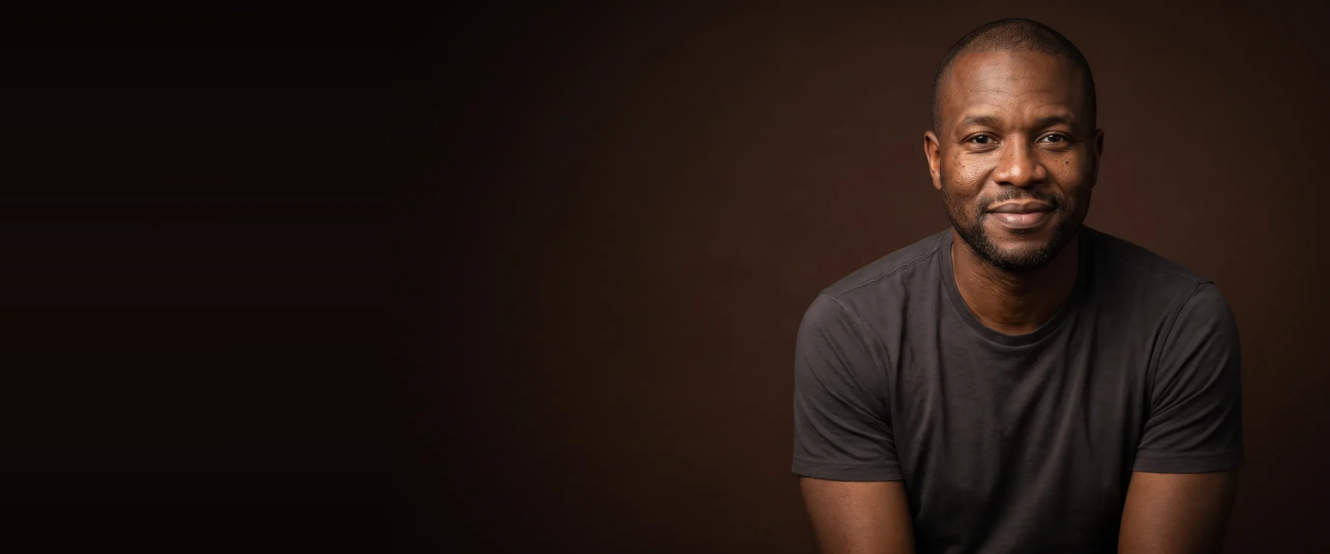 A man with a shaved head and short beard, holding back a smile, on a deep brown background