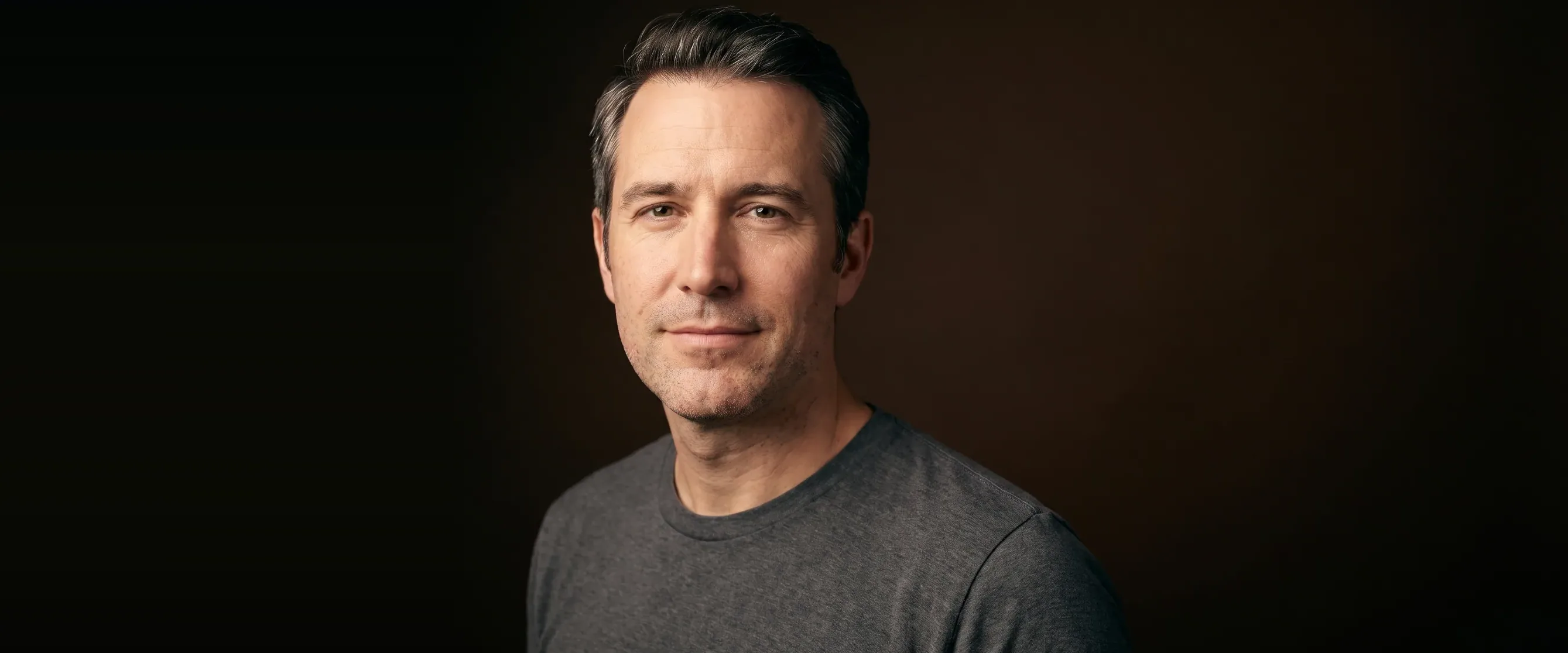 A man with greying temples, closely shaved, holding back a smile, on a deep brown background