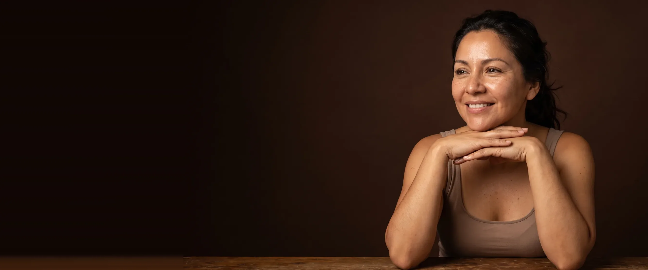 A woman resting her chin on her hands, a quiet smile, luminous skin