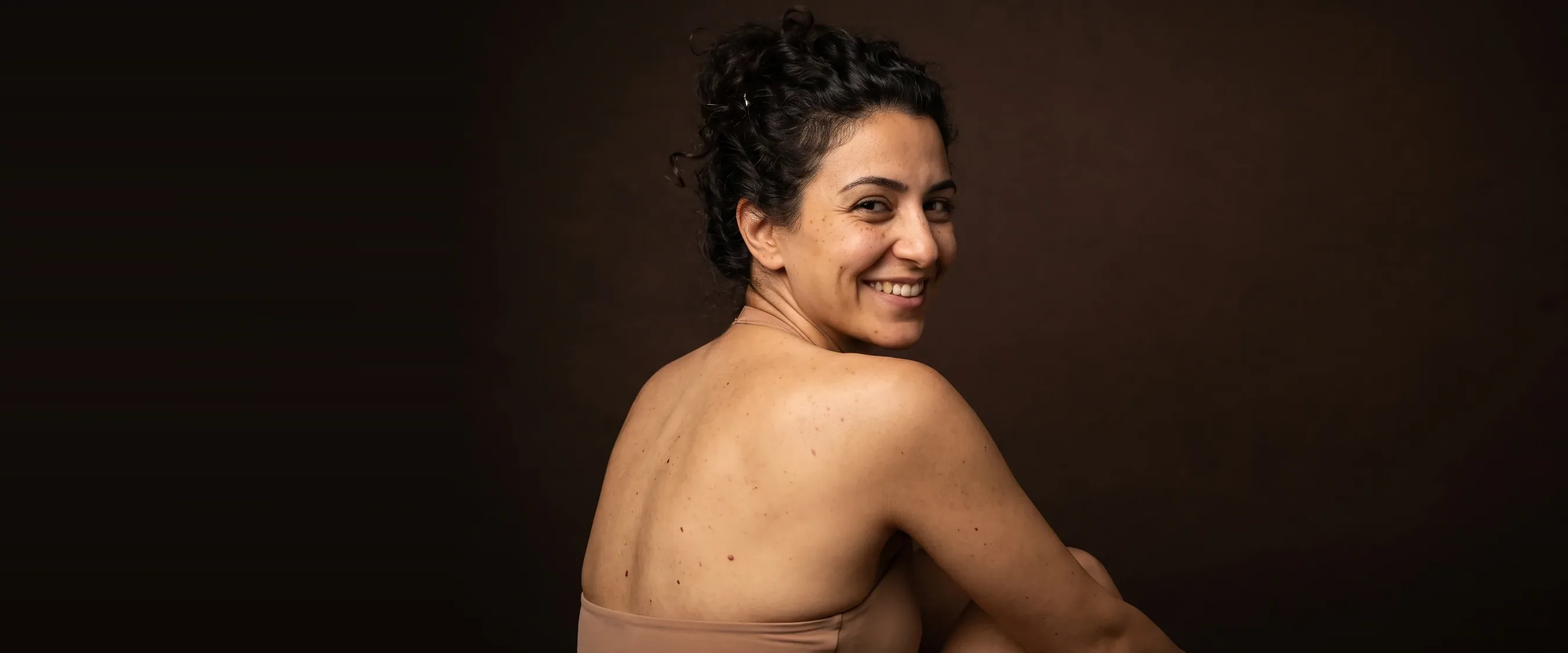 A woman seen from behind, shoulders bare, turned to the camera with a broad smile